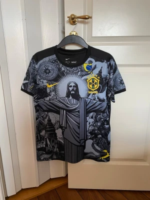 Nike Brasil svart t-shirt  - Cool svart t-shirt från Nike med Brasil-logga och unikt grafiskt tryck på framsidan som visar Kristusstatyn och detaljerade mönster. Gult och blått inslag i loggor och tryck. Perfekt för sport och träning.