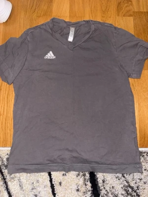 Grå v-ringad t-shirt  - Grå t-shirt från Adidas med v-ringning och korta ärmar. Diskret Adidas-logga på bröstet. Tillverkad i mjuk bomull som känns skön mot huden. Perfekt för dig som gillar sportiga och stilrena plagg.
