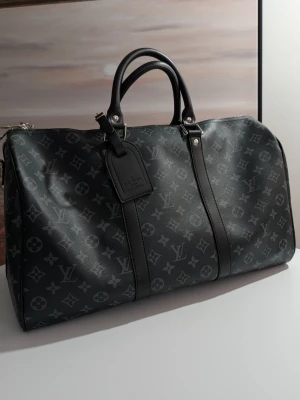 Iouis Vuitton duffelväska - Detta är en klassisk duffelväska (eller weekendväska) med en rymlig, cylinderformad kropp och rundade gavlar. Väskan är tillverkad i ett mörkt, belagt canvasmaterial med ett återkommande grafiskt mönster bestående av stiliserade blommor och geometriska symboler i en ljusgrå nyans mot en svart bakgrund.Väskan kännetecknas av en monokrom design där svarta läderdetaljer och silverfärgad metallhårdvara möter den mönstrade ytan. 