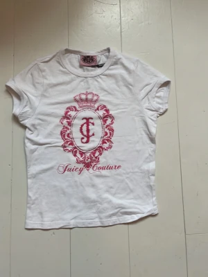 Vit Juicy Couture t-shirt med tryck - Snygg vit t-shirt från Juicy Couture med rosa emblemtryck och krona framtill. Klassisk rund halsringning och korta ärmar. Tillverkad i mjuk bomull som känns skön mot huden. Perfekt för dig som gillar en clean men ändå statement look.