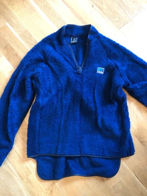 Blå fleece från Helly Hansen - Klassisk blå fleece från Helly Hansen med halv dragkedja och broderad logga på bröstet. Långärmad. Storlek small (men’s 46-48, ladies 40-42). Fint skick. 