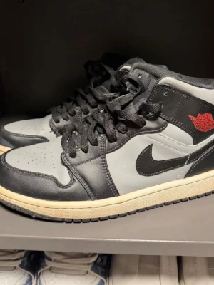 Nike Air Jordan 1 Mid svart/grå - Säljer ett par Nike Air Jordan 1 Mid sneakers i svart och grått med röda detaljer. Skorna har klassisk hög siluett, svart swoosh och snörning. Ovandelen är i läder och yttersulan är beige. Perfekta för dig som gillar streetwear och ikoniska sneakers.