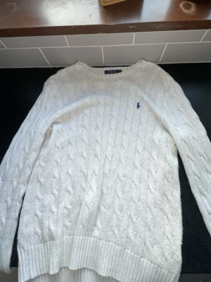 Vit kabelstickad tröja Polo Ralph Lauren - Kabelstickad vit tröja från Polo Ralph Lauren med klassisk blå logga broderad på bröstet. Tröjan har rund halsringning, ribbade muddar och är långärmad. Perfekt för dig som gillar tidlös och clean stil.