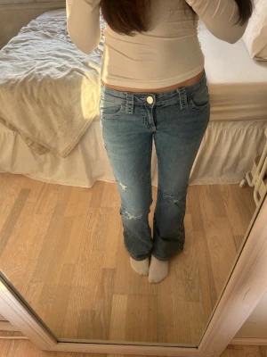 Bootcut jeans med broderad ficka - Säljer ett par ljusblå bootcut jeans med slitningar på knäna och snygg broderad detalj på bakfickan. Jeansen har låg midja och klassisk femficksdesign. Perfekta för dig som gillar en avslappnad och trendig stil.