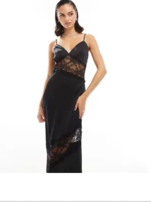 Svart långklänning med spetsdetaljer - Svart långklänning med tunna axelband och v-ringad urringning. Klänningen har genomskinliga spetsdetaljer vid midjan och nedtill, vilket ger en elegant och trendig look. Perfekt för dig som vill sticka ut med en stilren och modern outfit.
