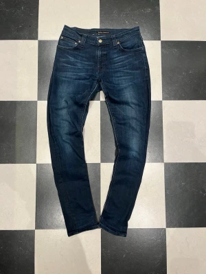 Mörkblå slim fit jeans från Nudie - Snygga mörkblå slim fit jeans från Nudie Jeans med klassisk femficksdesign och kontrastsömmar. Jeansen har en lätt tvättad look och är tillverkade i stretchigt denimtyg för extra komfort. Perfekta för en avslappnad och trendig stil.