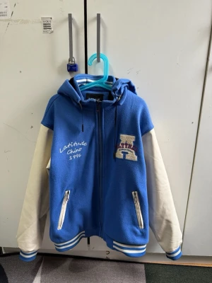 Blå och vit hoodie med brodyr - Snygg blå hoodie med vita ärmar och broderad text 'Latitude Chino 1996' på bröstet. Har dragkedja, två fickor med dragkedja och en stor huva. Detaljer i vitt och blått på muddar och nederkant. Perfekt för en sportig och avslappnad stil. För 10-12 åringar!