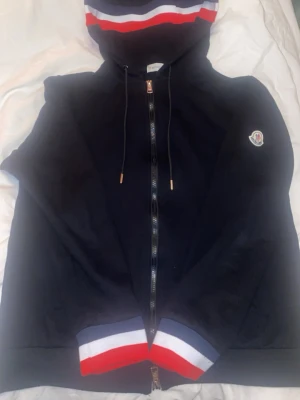 Svart Moncler hoodie med dragkedja - Svart hoodie från Moncler med hel dragkedja, huva och ribbade muddar. Snygga röd, vit och blå ränder på huva och ärmslut. Moncler-logga på vänster ärm. Perfekt för en stilren och sportig look.