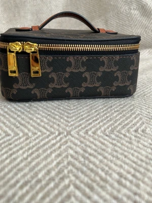  Triomphe monogram handväska - Snygg handväska sminkväska Triomphe-monogram i brunt och mörkbrunt canvasmaterial. Väskan har guldfärgade dragkedjor och ett brunt handtag i läder. Perfekt för dig som gillar lyxiga accessoarer med ikonisk design.