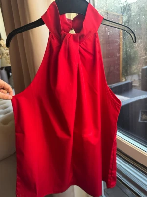 Röd halterneck topp med knyt - Snygg röd halterneck topp med knyt i halsen. Toppen är ärmlös och har en lös passform som ger en avslappnad men ändå chic look. Perfekt att styla med jeans eller kjol för en trendig outfit.