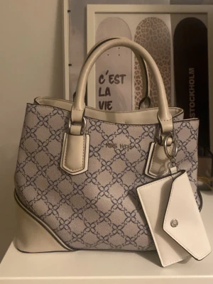 Mönstrad handväska från Nine West - Snygg handväska från Nine West i beige och ljusgrått med mörkblått mönster. Väskan har två korta handtag, dekorativ tag och flera innerfack, inklusive ett med dragkedja. Tillverkad i konstläder med silverfärgade detaljer. Perfekt för dig som gillar stilrena accessoarer. Orginal pris 1200kr