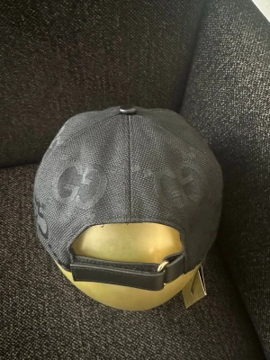 Svart keps från Gucci med GG-mönster - Snygg svart keps från Gucci med det klassiska GG-mönstret över hela kepsen. Justerbar rem bak i läder och böjd skärm med läderdetaljer. Perfekt accessoar för dig som vill ha en exklusiv och trendig look.