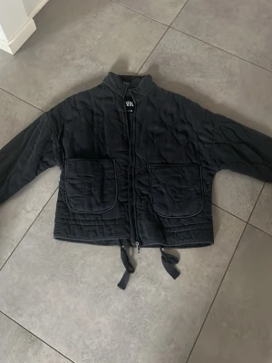 Svart quiltad jacka från Zara - Säljer en svart quiltad jacka från Zara med två stora fickor framtill och dragkedja. Jackan har knytband nedtill och en avslappnad passform. Perfekt för lager på lager och ger en cool, trendig look till din outfit.