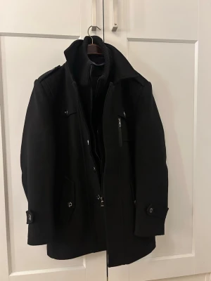 Svart klassisk ullkappa herr - Stilren svart ullkappa med hög krage och dragkedja samt knappar. Kappan har flera fickor, axelklaffar och detaljerade manschetter. Perfekt för kyliga dagar och ger en snygg siluett. Fodrad insida för extra komfort.