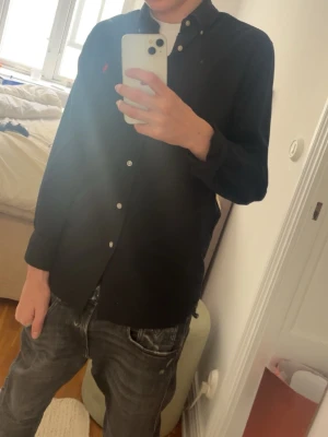 Svart skjorta från Ralph Lauren - Klassisk svart skjorta från Ralph Lauren med knappkrage och den ikoniska röda loggan broderad på bröstet. Skjortan har vita knappar och är långärmad. Tillverkad i bomull för en bekväm känsla och snygg passform.