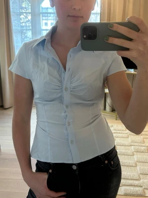 Ljusblå figursydd skjorta med knappar - Säljer en ljusblå figursydd skjorta med korta ärmar och klassisk krage. Skjortan har knappar framtill, rynkade detaljer vid bysten och knytband i ryggen för extra passform. Perfekt till jeans eller kjol för en clean och fräsch look.