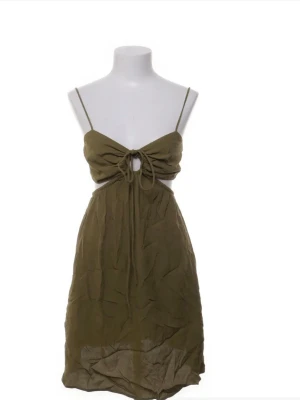 Olivgrön kort klänning med knyt - Snygg olivgrön kort klänning med smala axelband och knyt framtill. Klänningen har en liten cut-out under bysten och öppen rygg med resår. Perfekt för varma dagar och sommarens alla äventyr.