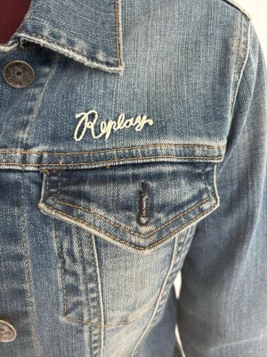 Blå jeansjacka från Replay - Snygg blå jeansjacka från Replay med klassisk krage, knappstängning och två bröstfickor med lock. Jackan har vit broderad Replay-logga på bröstet och kontrastsömmar. Figurnära passform och tillverkad i slitstarkt denimtyg.