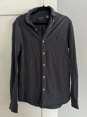 Mörkgrå skjorta från Polo Ralph Lauren - Mörkgrå skjorta från Polo Ralph Lauren med klassisk krage och vita knappar framtill. Skjortan har lång ärm och diskret broderad logga på bröstet. Tillverkad i mjuk bomull för en bekväm känsla och stilren look. Passar till mycket och är knappt använd! Nypris 999