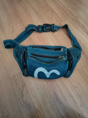 Evisu Blå bröst väska i denim med vit detalj - Säljer en blå bröst väska i denim med 6 dragkedjefack och justerbart spänne. Väskan har en vit målad detalj på framsidan och gula kontrastsömmar. Perfekt för att bära det viktigaste och ger en cool streetstyle vibe.