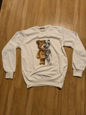 Vit sweatshirt med björn- och robottryck - Cool vit sweatshirt med ett unikt tryck av en nallebjörn som är halvt robot på framsidan. Tröjan har rund hals, långa ärmar och ribbade muddar vid ärmslut och nederkant. Perfekt för dig som gillar streetwear och statement-plagg.