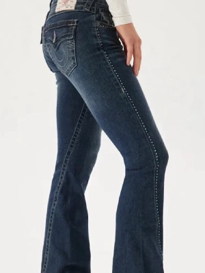 Blå bootcut jeans med nitar True Religion - Mörkblå bootcut jeans från True Religion med dekorativa nitar längs sidsömmarna. Jeansen är i modellen ”Joey crystal” och är helt slutsålda och går inte att köpa längre. Därav är de väldigt eftertraktade.