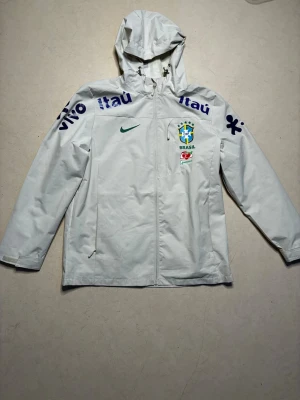 Vit Brasil Nike vindjacka med huva - Snygg vit vindjacka från Nike med Brasil-logga och sponsormärken som Itau och Vivo. Jackan har huva med dragsko, hel dragkedja framtill och två sidofickor. Perfekt för dig som gillar sportig stil och vill sticka ut med landslagskänsla.
