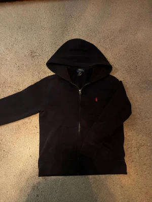 mörk blå  hoodie från Polo Ralph Lauren - Klassisk svart hoodie från Polo Ralph Lauren med dragkedja framtill och huva. Den har två fickor och den ikoniska röda loggan broderad på bröstet. Tillverkad i mjukt bomullsmaterial som är skönt att ha på sig. Perfekt för en avslappnad och stilren look.