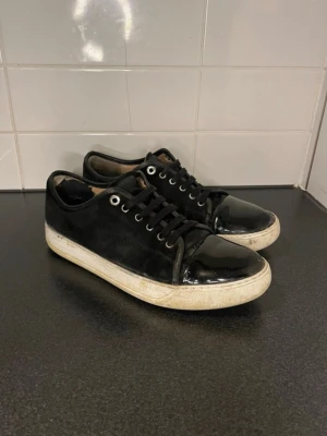  Lanvin - Snygga svarta sneakers från Lanvin med klassisk låg profil. Skorna har en mix av mocka och glansiga lackdetaljer på tån och hälen, samt vita sulor med tydlig Lanvin-logga undertill. Perfekta för dig som gillar stilrena och exklusiva sneakers.