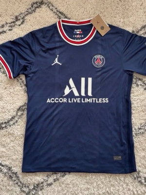 PSG matchtröja blå Jordan x Nike - Mörkblå PSG matchtröja med vita och röda detaljer i krage och ärmslut. Tröjan har Paris Saint-Germain-logga på bröstet, Jordan-logga och sponsortryck framtill. Tillverkad i lätt och ventilerande material, perfekt för fotboll eller träning.