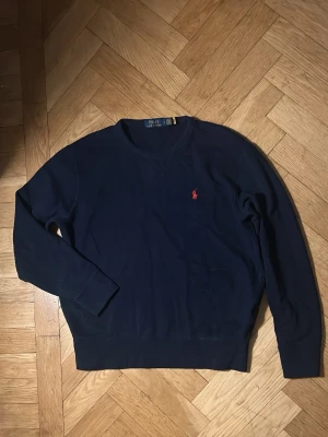 Mörkblå sweatshirt från Polo Ralph Lauren - Klassisk mörkblå sweatshirt från Polo Ralph Lauren med rund halsringning och den ikoniska röda logotypen broderad på bröstet. Tröjan har ribbade muddar vid ärmslut och nederkant, perfekt för en stilren och avslappnad look. Knappt använd. Original pris 2000kr