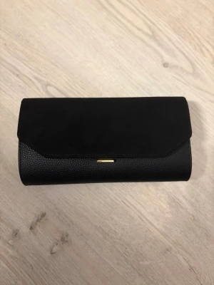 Svart kuvertväska med gulddetaljer - ✨perfekt till fest & speciella tillfällen! ✨  Nu säljer jag en superfin kuvertväska som verkligen lyfter din outfit 💫   Locket är i mjukt, lyxigt sametsmaterial som ger en exklusiv känsla, medan resten av väskan är i stilrent skinn – en perfekt kombination av klassiskt och trendigt.  🖤 Endast använd 1 gång – i princip som ny   🖤 Perfekt storlek för mobil, kort, smink m.m.   🖤 Passar både till fest, bröllop, student eller middagar 