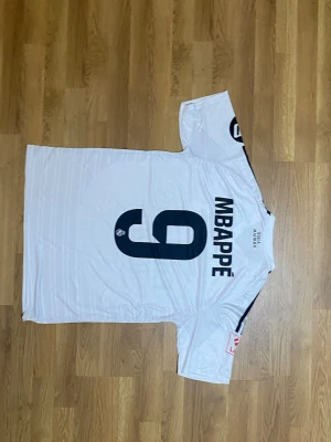 Real Madrid Mbappé matchtröja vit - Säljer en vit Real Madrid matchtröja från Adidas med Mbappé och nummer 9 på ryggen. Tröjan har svarta detaljer, klubbmärke och sponsortryck på framsidan samt klassiska Adidas-ränder på axlarna. Perfekt för fotbollsfans!