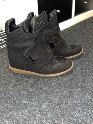 Svarta wedge sneakers med kardborre - Snygga svarta sneakers med inbyggd kilklack och tre breda kardborreband. Skorna har en mix av mocka och textil, samt detaljer i läderimitation. Yttersulan är i gummi med brun färg. Perfekta för dig som vill ha lite extra höjd och streetstyle.