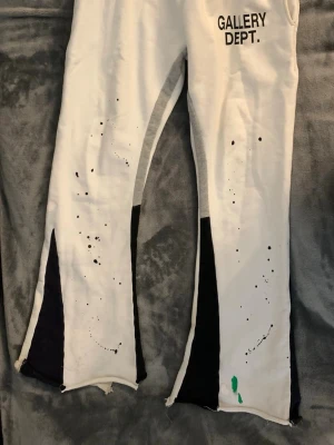 Vita mjukisbyxor Gallery Dept. - Unika vita mjukisbyxor från Gallery Dept. med svart text på höften, färgstänk och slitna detaljer. Byxorna har svarta paneler längs benen och råa kanter vid bensluten. Tillverkade i mjukt material för maximal komfort och streetwear-vibe.