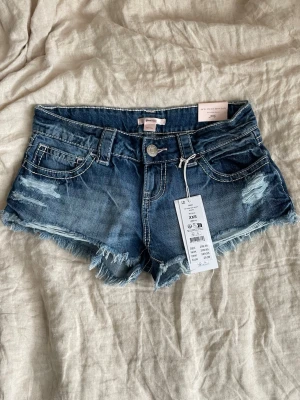 Blå jeansshorts  - Säljer ett par blå denimshorts med fransig kant och slitna detaljer. Klassisk femficksmodell med låg midja och råa kanter för en avslappnad look. Perfekta till sommaren och lätta att matcha med olika toppar. Skriv om ni har andra frågor!🥰