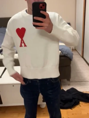 Vit stickad tröja från Ami Paris - Vit stickad tröja från Ami Paris med röd hjärtlogga och bokstaven A på bröstet. Tröjan har rund hals, ribbade muddar och avslut. Perfekt oversized fit och mjukt material som känns lyxigt.