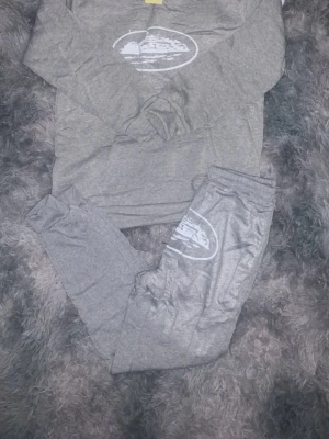 Grått corteiz  loungewear-set  - Mysigt grått loungewear-set med hoodie och byxor. Setet har ett vitt tryck med motiv på både tröjan och byxorna. Hoodien har känguruficka och dragsko i huvan. Perfekt för chill dagar hemma eller när du vill vara bekväm.