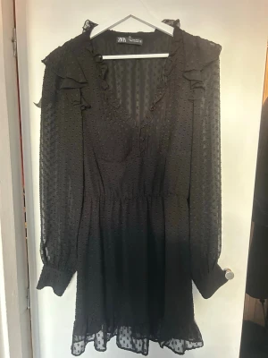 Svart kort klänning från Zara - Säljer en svart kort klänning från Zara med v-ringning, volangdetaljer vid axlarna och transparenta långärmar. Klänningen har små prickar i tyget och resår i midjan för en snygg passform. Perfekt för dig som gillar feminina detaljer och lätt material.