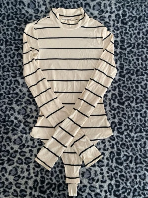 Randig långärmad polotopp - Säljer en beige och svart randig polotopp med lång ärm. Toppen har en smal passform och är ribbad i materialet, vilket ger en snygg struktur. Perfekt att styla med jeans eller kjol för en trendig look.