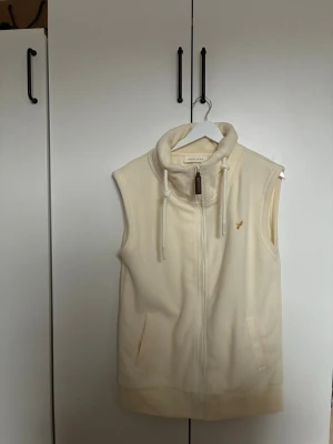 Krämvit fleeceväst med dragkedja från Pier one (OANVÄND) - Mysig krämvit fleeceväst från pier one med hög krage, dragsko och dragkedja framtill. Västen har två fickor och en liten gul logga på bröstet. Perfekt för lager på lager och chill stil.