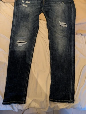 Mörkblå slitna jeans med raka ben - Säljer ett par mörkblå jeans med raka ben och slitna detaljer på låren och knäna. Jeansen har en klassisk femficksmodell och är tillverkade i denim med snygg tvätt och slitningar. Byxorna är ifrån jack and Jones i storlek w34 L32.