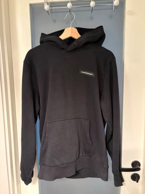 Svart hoodie från Peak Performance - Snygg svart hoodie från Peak Performance med klassisk känguruficka och liten logotyp på bröstet. Hoodien har en stor huva med dragsko och ribbade muddar vid ärmslut och nederkant. Perfekt för en avslappnad och stilren look.