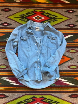 Ljusblå jeansskjorta från Acne - Säljer en klassisk ljusblå jeansskjorta från Acne med två bröstfickor och knappar framtill. Skjortan har lång ärm och en avslappnad passform, perfekt att styla lager på lager eller bära som den är.