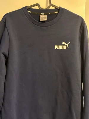 Mörkblå sweatshirt från Puma - Snygg mörkblå sweatshirt från Puma med klassisk logga i vitt på bröstet. Tröjan har rund halsringning och ribbade muddar vid ärmslut och nederkant. Perfekt för en avslappnad och sportig stil.