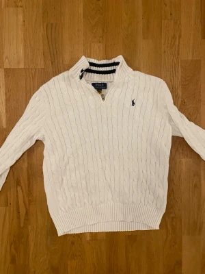 Vit kabelstickad tröja Polo Ralph Lauren - Kabelstickad vit tröja från Polo Ralph Lauren med halv dragkedja och hög krage. Tröjan har det klassiska broderade logotypen på bröstet och ribbade muddar vid ärmslut och nederkant. Perfekt för en stilren och avslappnad look.