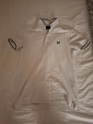 Vit pikétröja från Lyle & Scott - Klassisk vit pikétröja från Lyle & Scott med korta ärmar och svart piping på krage och ärmslut. Tröjan har knappslå framtill och den ikoniska gula loggan broderad på bröstet. Perfekt till en clean och sportig stil. Har dock några defekter vid armhålorna, men går att sy till om man vill.