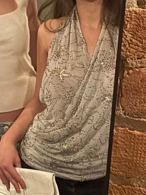 Glittrig omlott halterneck topp - Snygg glittrig halterneck topp i vitt med omlott-design och djup v-ringning. Toppen är ärmlös och har ett transparent, lätt material med silverfärgade paljetter som ger ett extra lyft till outfiten. Perfekt för dig som vill sticka ut.