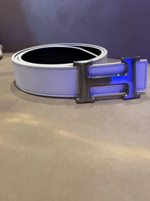 Vitt H’e’r’m’e”s skärp med H spänne - Snyggt vitt skärp från H’e’r’m’e”s med  H format spänne. Bältet och har en stilren, minimalistisk design som passar till många olika looks. Perfekt accessoar för dig som gillar exklusiva detaljer.