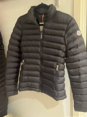 Svart dunjacka från Moncler - Snygg svart dunjacka från Moncler med quiltad design och hög krage. Jackan har två dragkedjeförsedda fickor framtill och en Moncler-logga på ärmen. Perfekt för kalla vinterdagar och riktigt stilren look.